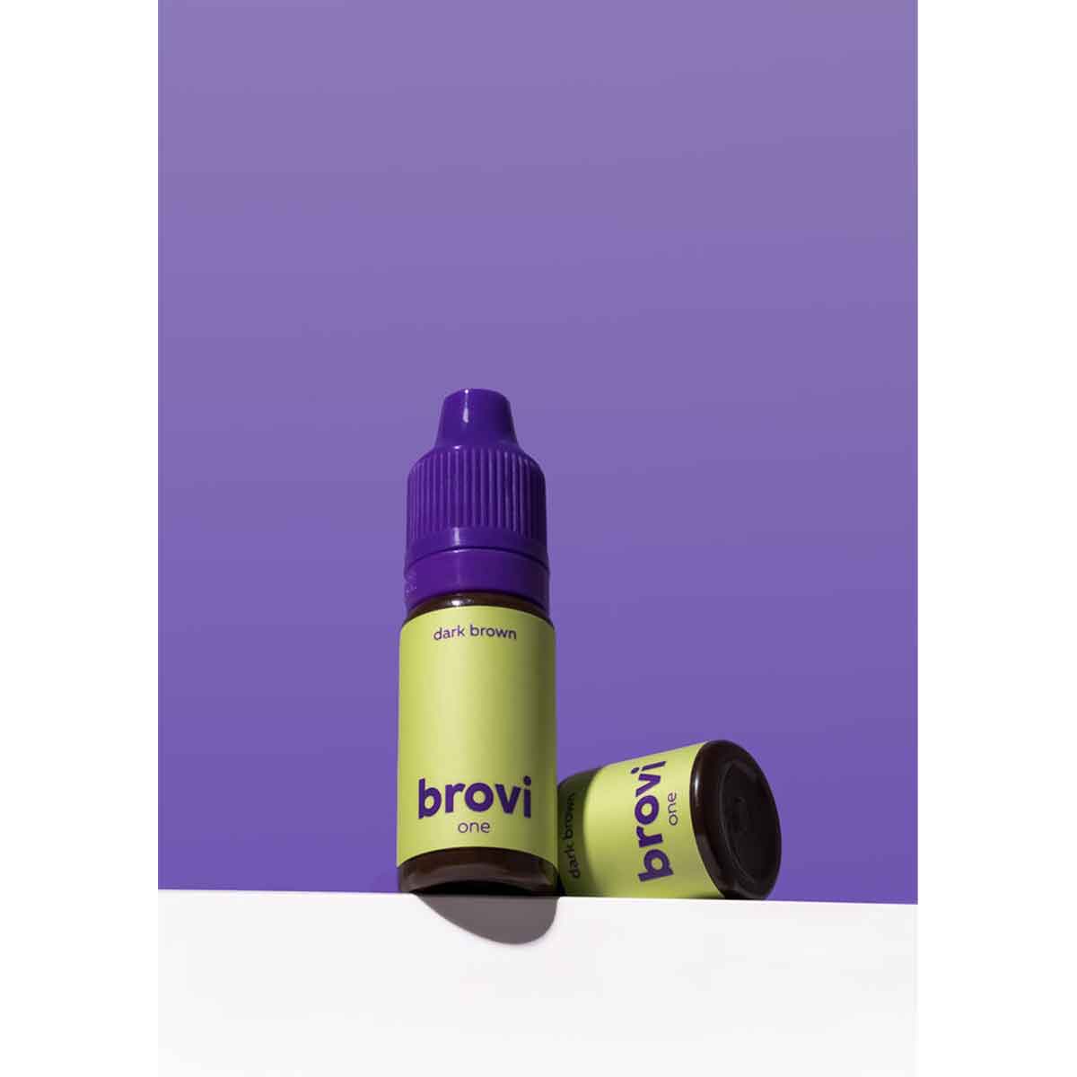 A bottle of Brovi One e-liquid in the 'dark brown' flavor, standing upright next to its cap on a white surface against a solid purple background.