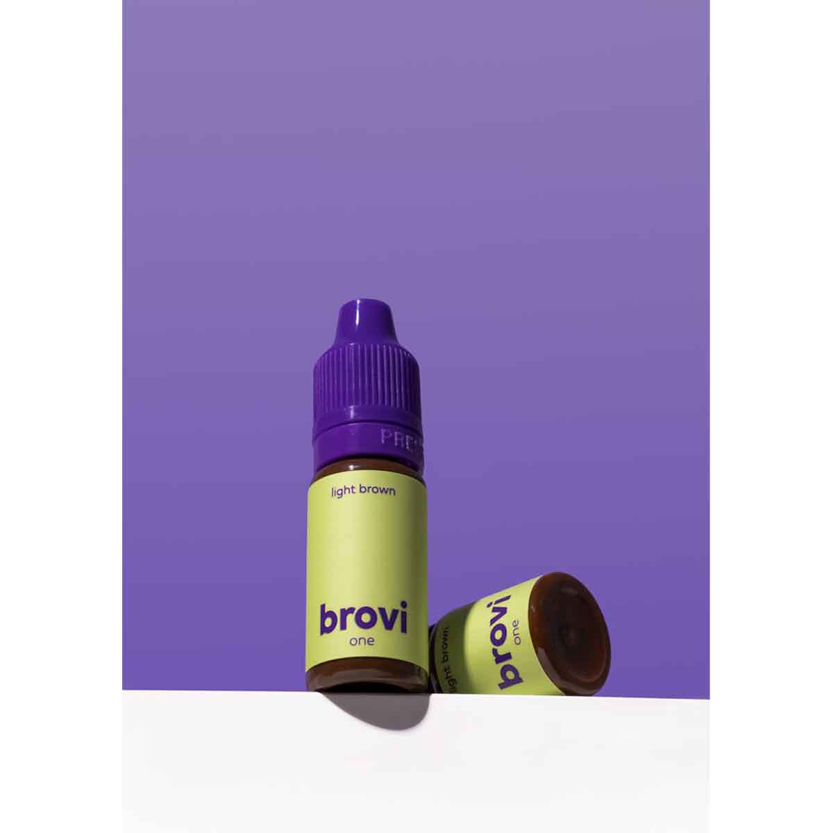 A bottle of Brovi One e-liquid in 'light brown' flavor, standing upright with its cap on, and another bottle lying on its side. The bottles have lime green labels and purple caps, set against a solid purple background with a white surface below.