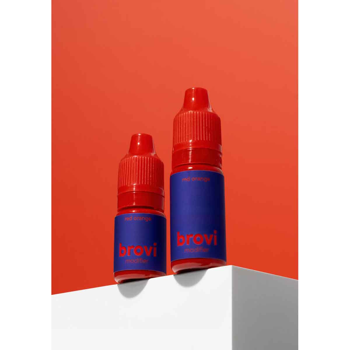 Two Brovi e-liquid bottles in 'mid orange' flavor, featuring blue labels and red caps, displayed on a white geometric pedestal against a solid orange-red background.