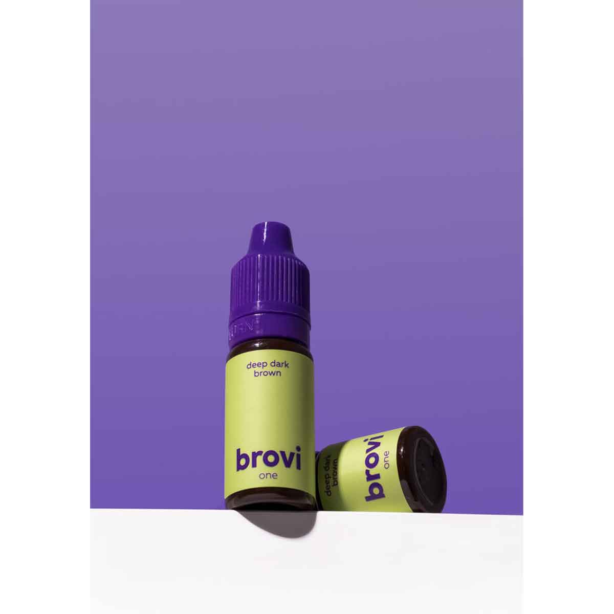 Two Brovi One e-liquid bottles in 'deep dark brown' flavor, featuring a light green label and purple caps, set against a solid purple background with a white surface.