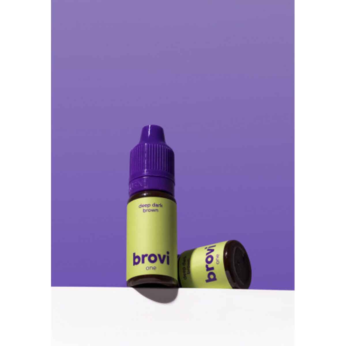 Two Brovi One e-liquid bottles in 'deep dark brown' flavor, one standing upright and the other lying on its side, set against a solid purple background with a white surface below.