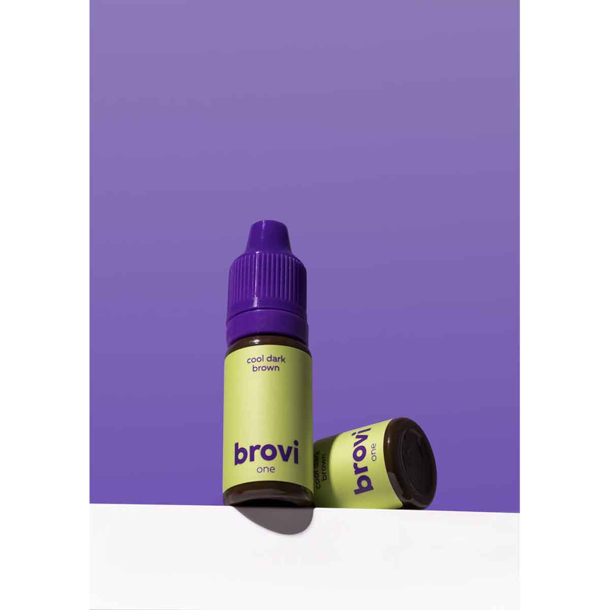 Brovi One coffee extract e-liquid bottle in 'cool dark brown' flavor, standing upright with another bottle lying on its side, set against a solid purple background and a white surface.