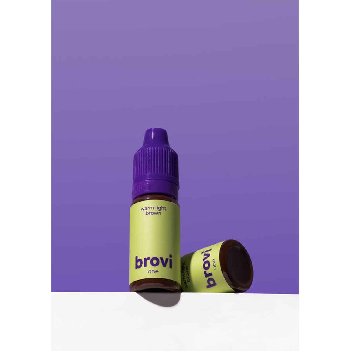 Brovi One e-liquid bottle in 'warm light brown' flavor, featuring a lime green label and purple cap, shown standing next to its matching purple cap on a white surface against a solid purple background.
