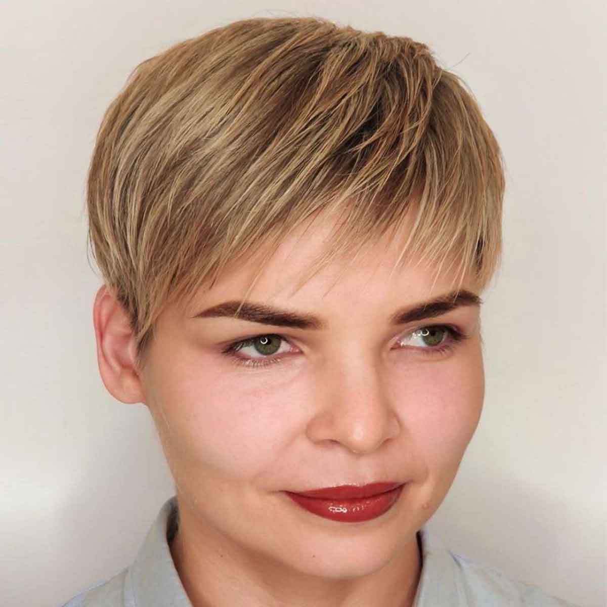 A close-up portrait of a person with a short, layered pixie cut hairstyle in light blonde and brown tones, wearing a light gray top against a plain off-white background.