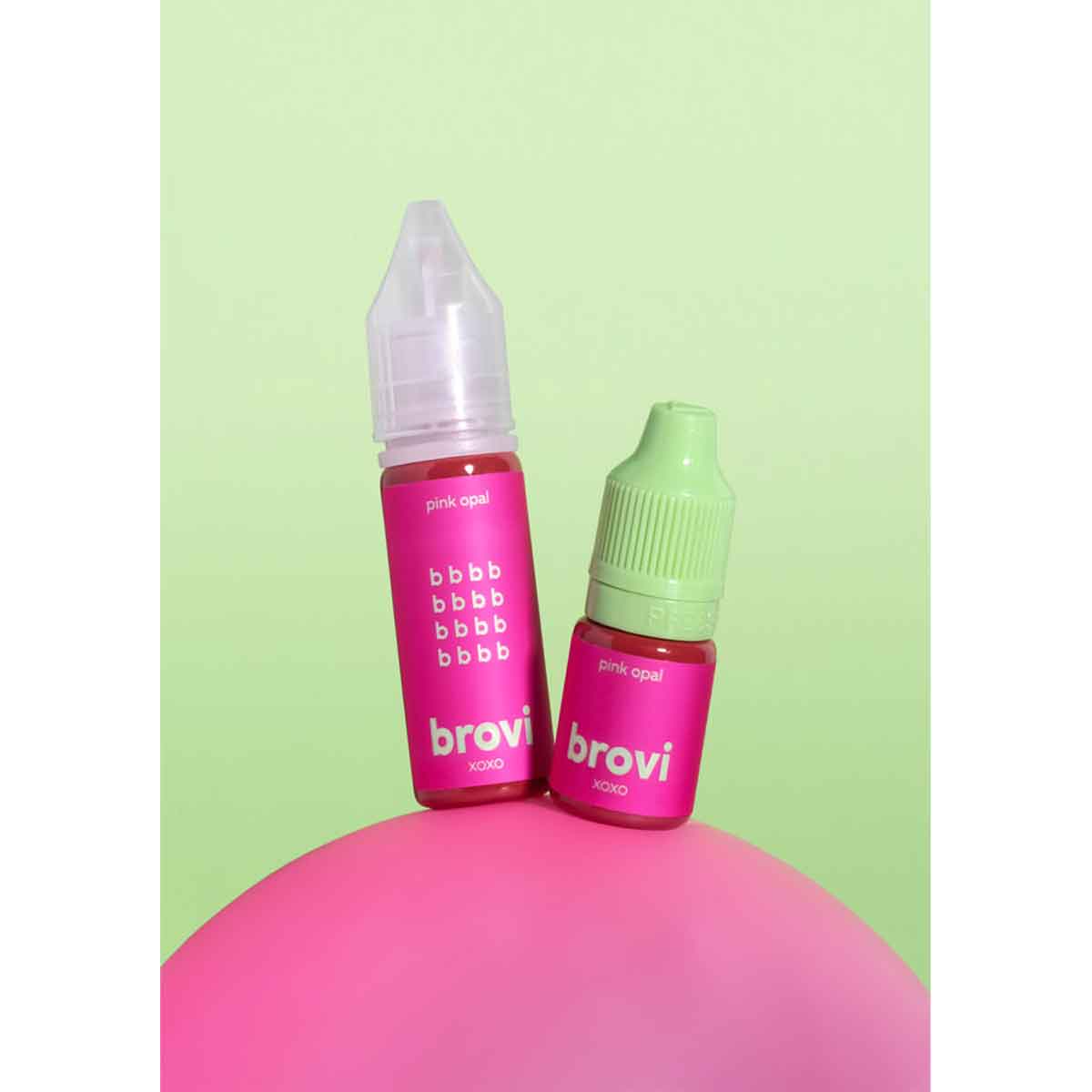 Two Brovi e-liquid bottles in the 'purrify liquid' flavor, one with a clear cap and one with a light green cap, both featuring bright pink labels, displayed on a pink sphere against a light green background.