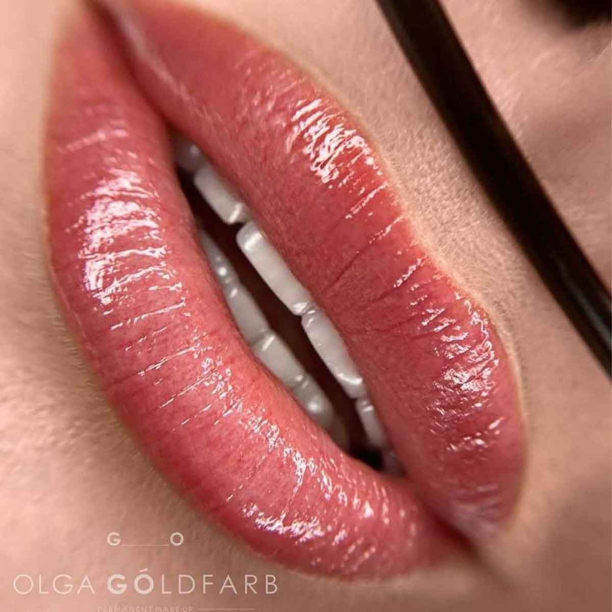 Close-up of glossy pink lips wearing a lip color, with part of a dark eyeglass frame visible on the right. The text 'OLGA GÖLDFARB' appears at the bottom left.