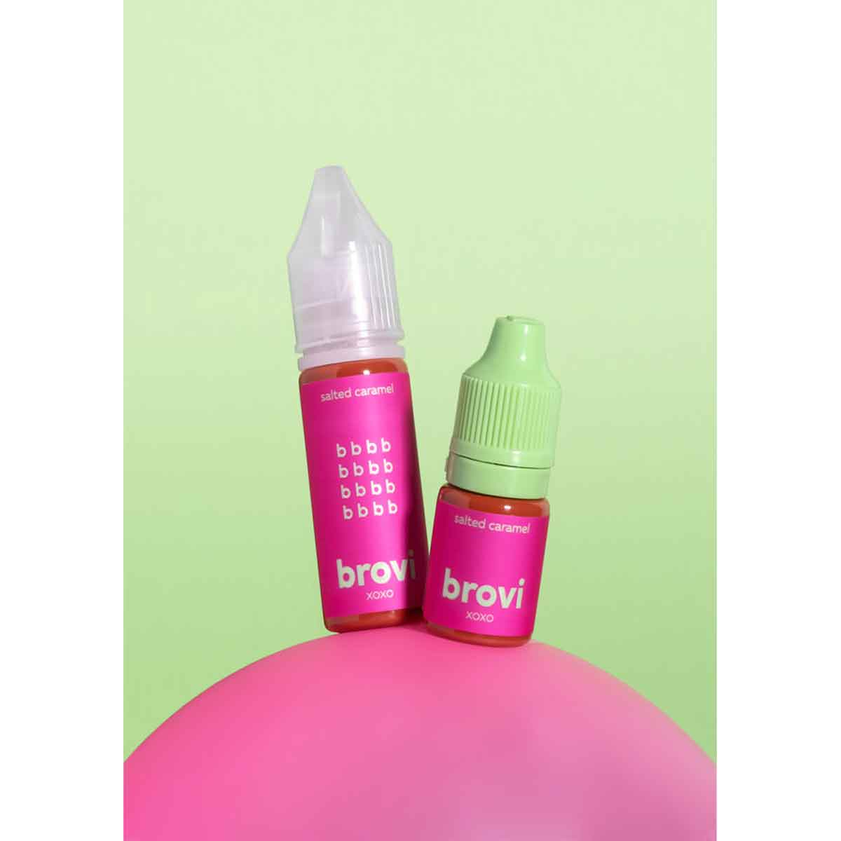 Two small Brovi e-liquid bottles on a pink rounded surface against a pale green background. One bottle has a white cap and the other has a light green cap, both with bright pink labels reading 'salted caramel' and 'brovi'.