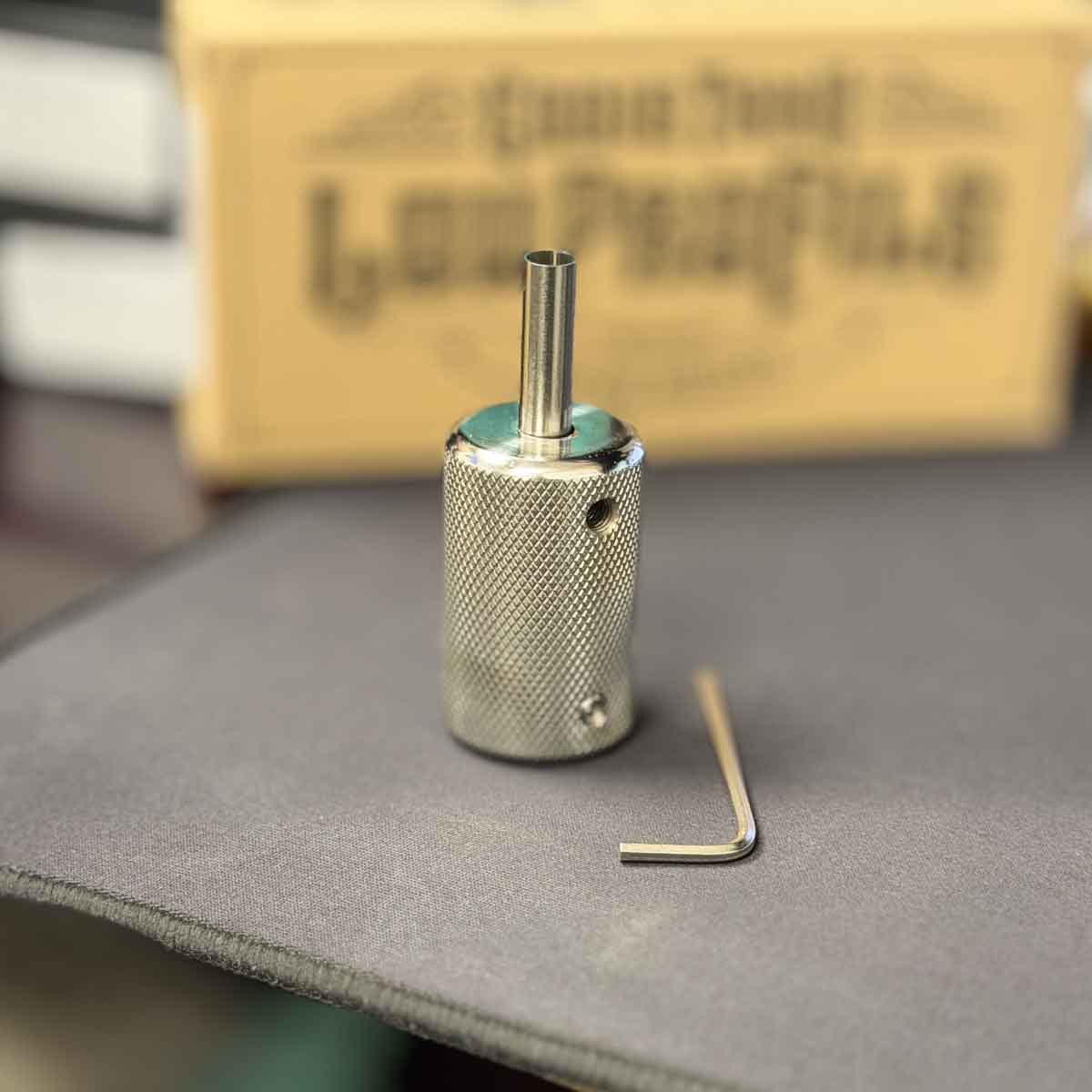 stainless steel tattoo grip with hex key on a gray surface, with a blurred background featuring text.