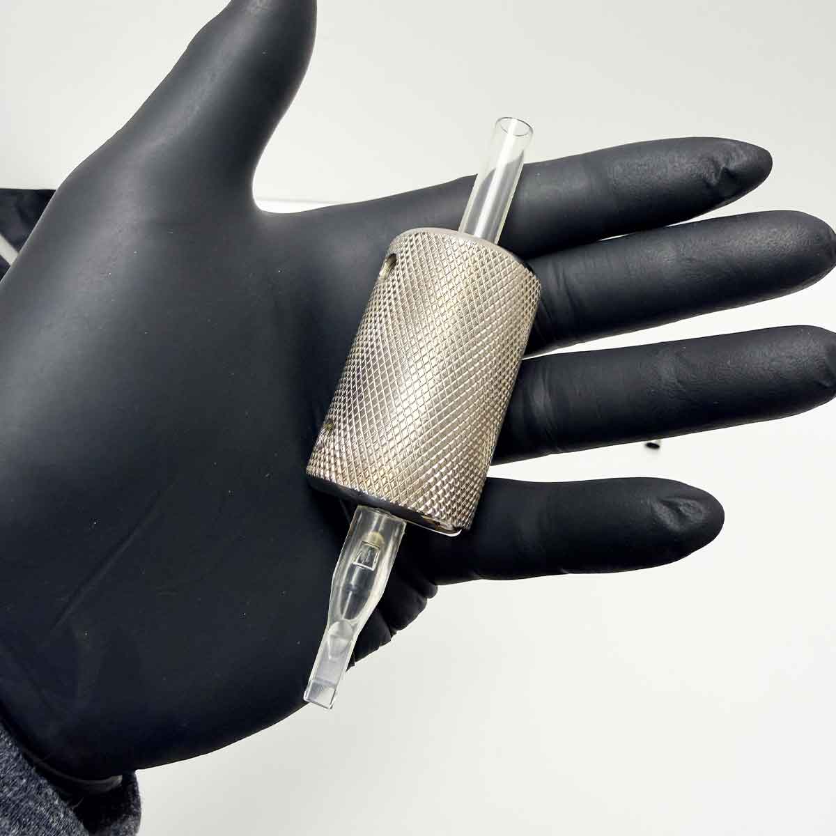 Hand wearing a black glove holding a stainless steel tattoo grip on a white background